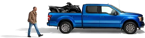 A man approaches his blue pickup truck with a black ATV in the bed.